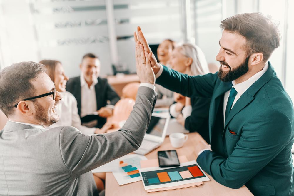 Two successful smiling architects in formal wear giving high five while sitting at board room. In background colleagues chatting. Average will not be celebrated.